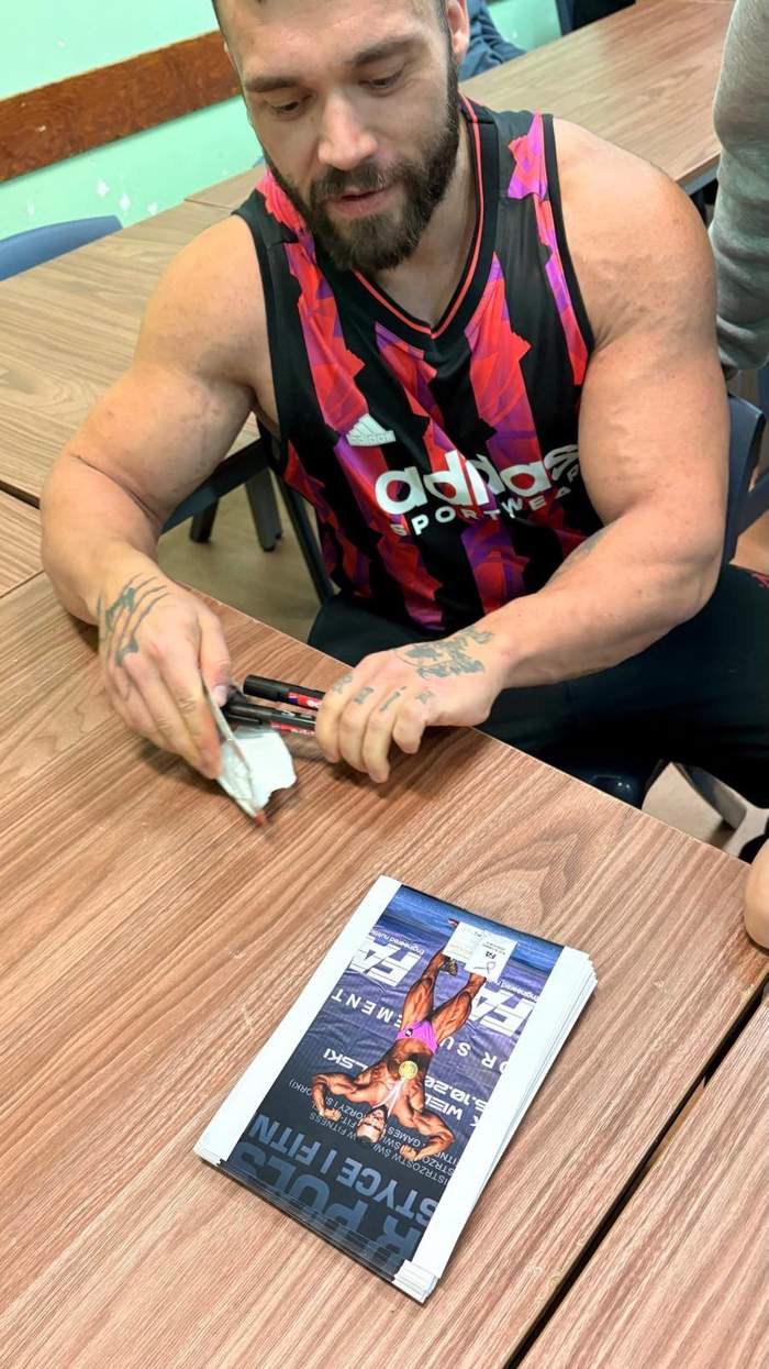 Pokaż zdjęcie: Silnie umięśniony mężczyzna w koszulce na ramiączkach rozdaje autografy na pamiątkowych zdjęciach