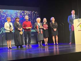 Pokaż zdjęcie: Na scenie Białostockiego Teatru Lalek stoi siedem osób: pięć kobiet i dwóch mężczyzn. Tomasz Klim Zastępca Prezydenta Miasta Białegostoku stojący po prawej stronie sceny przy mównicy składa gratulacje i podziękowania byłym i obecnym dyrektorom Zespołu Placówek Szkolno – Wychowawczych. Pierwsze cztery osoby stojące z lewej strony trzymają w rękach upominki.  Kobieta w czarne sukience pokazuje widowni list gratulacyjny w bordowej okładce.