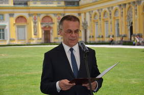 Pokaż zdjęcie: Mężczyzna w garniturze i okularach przemawia na stojąco, w tle zabudowania pałacowe