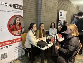 Pokaż zdjęcie: Za biurkiem siedzą trzy kobiety w marynarkach. Przed nimi siedzi kobieta w czarnej puchatej kurtce. Po lewej stronie stoi baner z tekstem w języku angielskim. Po prawej kolejne stanowiska. 