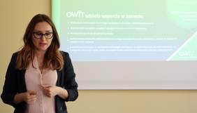 Pokaż zdjęcie: Kobieta w okularach znajduje się w sali konferencyjnej i wygłasza swoją prelekcję. W tle, na dużym ekranie wyświetlony jest slajd informujący o tym, jakie zadania pełnią Ośrodki Wsparcia i Testów.