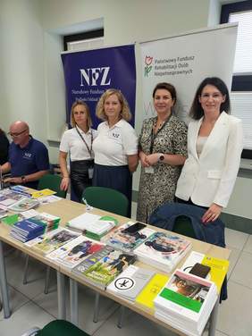 Pokaż zdjęcie: Na zdjęciu cztery kobiety pracownice NFZ i PFRON w Białymstoku stoją przy stole. Mężczyzna w niebieskiej koszulce z logo NFZ siedzi na końcu stół na którym leżą materiały informacyjne. Za nim stoją dwa banery z logo PFRON i NFZ.