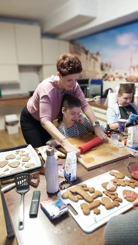 Pokaż zdjęcie: W kuchni znajduje się dwóch uczestników zajęć kulinarnych oraz kobieta, która pokazuje, w jaki sposób formuje się ciasto na pierniczki z wykorzystaniem drewnianego wałka. Na stole można dostrzec przygotowane różne rodzaje pierników.