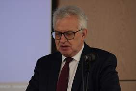 Pokaż zdjęcie: Przy mównicy na auli stoi mężczyzna z siwymi włosami i w okularach