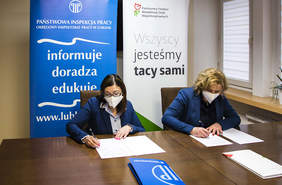 Pokaż zdjęcie: dwie kobiety w niebieskich marynarkach i maseczkach siedzą przy brązowym stole, przed nimi leżą dokumenty, które podpisują w tle loga PIP oraz PFRON