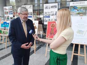 Pokaż zdjęcie: Dziennikarka przeprowadza wywiad z dyrektorem Łódzkiego Oddziału PFRON. W tle na sztalugach stoją prace artystyczne, które wygrały w Przeglądzie Twórczości Plastycznej Osób Chorych, Starszych i Niepełnosprawnych. 