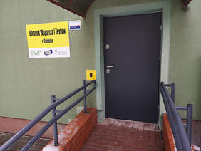 Pokaż zdjęcie: Wejście do budynku, szerokie brązowe drzwi, widać fragment zielonej elewacji, do drzwi prowadzi podjazd z metalowymi barierkami, po lewej stronie od wejścia tabliczka z napisem: Ośrodek Wsparcia i Testów w Świdniku, na tabliczce widać logo OWIT or