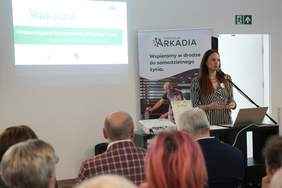 Grupa osób, siedzą, kobieta stoi za mównicą, w tle kolorowy baner