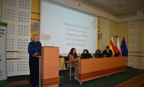 Pokaż zdjęcie: Na zdjęciu sala konferencyjna z podium i stołem prezydialnym. Kobieta w granatowej bluzce i białych okularach, stoi przy mównicy i przemawia do uczestników. Za nią na ekranie wyświetlana jest prezentacja o wykorzystaniu środków z budżetu państwa i PFRON na placówki wsparcia. Przy stole siedzą cztery kobiety, ubrane w formalne stroje, korzystające z laptopów. W tle widoczne są flagi Polski, województwa podlaskiego i Unii Europejskiej.