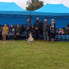 Pokaż zdjęcie: Na dworze pod namiotem siedzą na ławkach osoby, przed namiotem stoją w rzędzie cztery osoby, przy jednym z mężczyzn stoi kosz z cukierkami