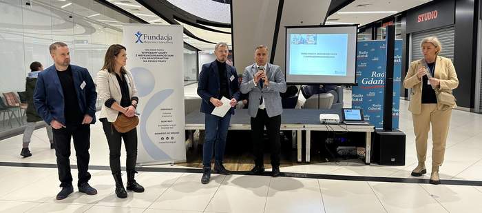 Pokaż zdjęcie: Koło siebie stoi dwóch mężczyzn w marynarkach, jden trzyma mikrofon. Po prawej stoi kobieta w jasnym garniturze która miga. Po lewej baner z logo Fundacji i kobieta z mężczyzną. Obydwoje w marynarkach. Za nimi widać piętro galerii i schody rucho