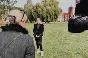 Pokaż zdjęcie: Pośrodku zdjęcia stoi na trawie młoda dziewczyna, która się wypowiada. Po prawej stronie widać obiektyw kamery, a po lewej stronie rozmwiającą z nią dziennikarkę. 