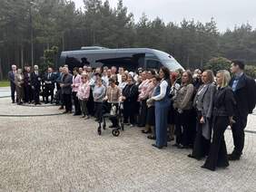 Pokaż zdjęcie: Grupa kilkudziesięciu osób stoi przed autobusem zaparkowanym na placu wyłożonym kostką brukową. W tle znajduje się las iglasty. 