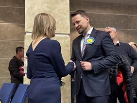 Pokaż zdjęcie: Zbliżenie na dwoje ludzi podczas oficjalnego powitania lub gratulacji. Mężczyzna w ciemnym, prążkowanym garniturze z żółtą przypinką "słoneczkiem" uśmiecha się i podaje rękę kobiecie w granatowej sukience, która stoi tyłem do obiektywu. Scena rozgrywa się przy jasnym filarze w nowoczesnym wnętrzu. W tle widać inne osoby uczestniczące w wydarzeniu.