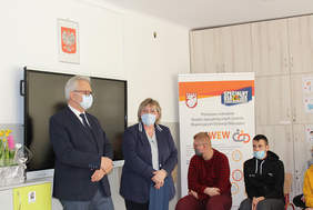 Pokaż zdjęcie: Na tle czarnej tablicy stoi mężczyzna w garniturze oraz kobieta, w tle dzieci