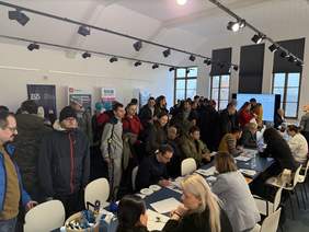 Pokaż zdjęcie: Tłum ludzi w jasnym pomieszczeniu. Kilka osób siedzi przy stołach, rozmawiając i wypełniając dokumenty, a inni stoją w kolejce. W tle widoczne są banery firm i instytucji.