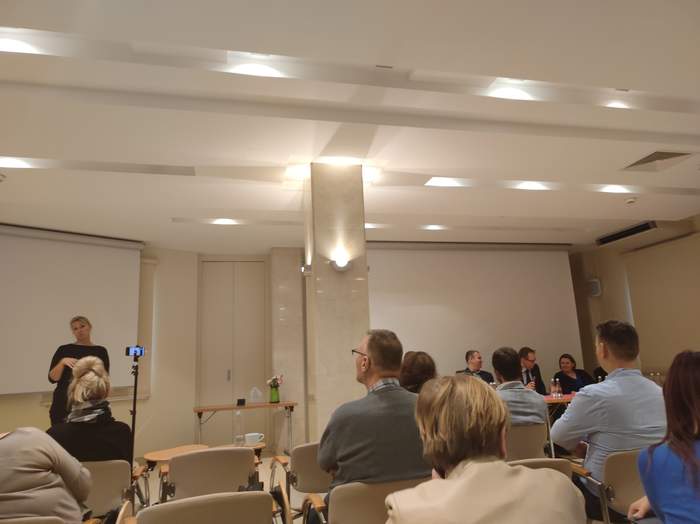 Ludzie siedzący na sali, na przeciwko nich za stołem siedzą osoby prowadzące konferencję. W lewym roku stoi kobieta - tłumacz języka migowego.