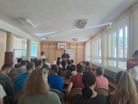 Pokaż zdjęcie: Zdjęcie uczestników spotkania siedzących na krzesełkach, z przodu sali stoją prelegenci kobieta i mężczyzna.