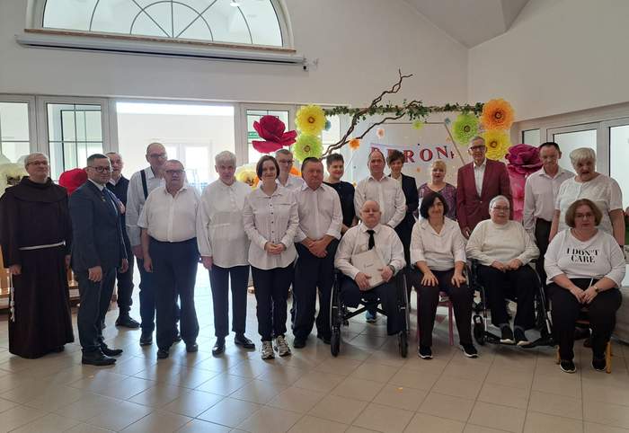 Pokaż zdjęcie: Grupa osób w eleganckich strojach pozuje w dwóch rzędach w jasnym pomieszczeniu. W tle dekoracje z dużych papierowych kwiatów i napis „PFRON”. Jeden z mężczyzn ma na sobie charakterystyczną czerwoną marynarkę.