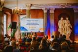 Sala konferencyjna z publicznością siedzącą na krzesłach i zwróconą w stronę sceny. Na środku widać mównicę. Po lewej stronie sceny ustawiony jest ekran projekcyjny i wyświetlony jest na nim slajd. Za mównicą znajdują się wysokie kolumny oraz duża jasna rzeźba przedstawiająca dwie stojące postacie ludzkie na postumencie.  Ściany mają ciepły, czerwony kolor, a oświetlenie w odcieniach pomarańczowo niebieskich podkreśla kolumny i rzeźbę.  Tekst na ekranie projekcyjnym: „Serdecznie Witamy Państwa na spotkaniu zespołu Specjalistycznego Centrum Wspierającego Edukację Włączającą poniżej napis SCWEW  i data 7 maja 2024.