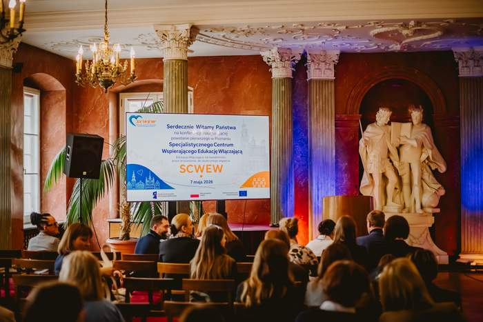 Pokaż zdjęcie: Sala konferencyjna z publicznością siedzącą na krzesłach i zwróconą w stronę sceny. Na środku widać mównicę. Po lewej stronie sceny ustawiony jest ekran projekcyjny i wyświetlony jest na nim slajd. Za mównicą znajdują się wysokie kolumny oraz duża jasna rzeźba przedstawiająca dwie stojące postacie ludzkie na postumencie.  Ściany mają ciepły, czerwony kolor, a oświetlenie w odcieniach pomarańczowo niebieskich podkreśla kolumny i rzeźbę.  Tekst na ekranie projekcyjnym: „Serdecznie Witamy Państwa na spotkaniu zespołu Specjalistycznego Centrum Wspierającego Edukację Włączającą poniżej napis SCWEW  i data 7 maja 2024.
