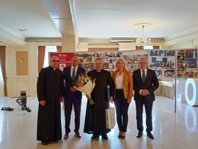 5 osób, ksiądz w sutannie i ciemnych okularach, następnie kobieta w rudej marynarce, spodniach obok niej mężczyzna w garniturze mężczyzna w garniturze, drugi ksiądz, trzyma bukiet kwiatów, 