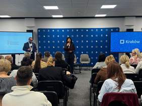 Pokaż zdjęcie: Sala konferencyjna. Na pierwszym planie uczestnicy siedzący na krzesłach słuchają prowadzących. Prowadzący kobieta i mężczyzna przemawiają do mikrofonów. 