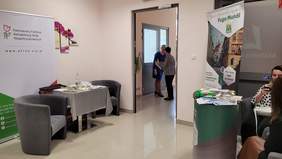 Pokaż zdjęcie: Na korytarzu po lewej stronie w kącie stoi rollup z logo PFRON, obok stolik z ulotkami oraz dwa fotele. Po drugiej stronie korytarza stoisko informacyjne FUGA MUNDI. 