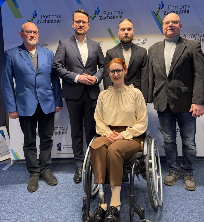 Pokaż zdjęcie: Portretowe zdjęcie pięciu osób pozujących na tle ścianki z logo „Pomorze Zachodnie”. W centrum siedzi uśmiechnięta kobieta w okularach na wózku inwalidzkim, ubrana w beżową bluzkę z falbaną przy szyi i brązowe spodnie. Za nią stoi czterech mężczyzn w marynarkach. Całość kompozycji jest jasna i oficjalna. Na dole ścianki widoczne są logotypy Funduszy Europejskich.