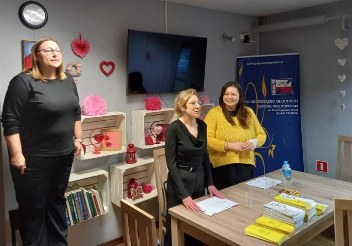 Pokaż zdjęcie: Dwie kobiety stoją za stolikiem, na którym wyłożone są materiały informacyjne. Po lewej stronie stoi kobieta, która tłumaczy spotkanie na język migowy. Po prawej stronie ustawiony jest baner Polskiego Związku Głuchych.