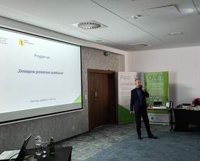 Pokaż zdjęcie: Na ekranie prezentacja, którą omawia pracownik PFRON, w tle stół ze sprzętem wspomagającym