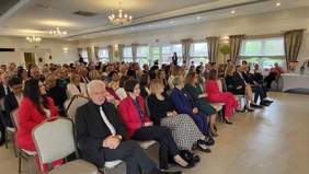 Pokaż zdjęcie: Grupa kilkudziesięciu osób siedzi w dużej sali konferencyjnej, na krzesłach ustawionych w kilku rzędach.