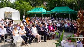 Pokaż zdjęcie: W plenerze w rzędach są ustawione krzesła. Siedzą na nich ludzie. Wszyscy są elegancko ubrani. Niektórzy trzymają na kolanach różne upominki, kwiaty i prezenty. W tle na markizach umieszczone są napisy: Dom Pomocy Spoełcznej w Wiśniowej Górze. 