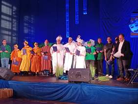 Pokaż zdjęcie: Na scenie stoi kilkanaście osób. Niektórzy mają na sobie przebrania, a inni garnitury i wytworne sukienki. Wspólnie śpiewają piosenkę. 