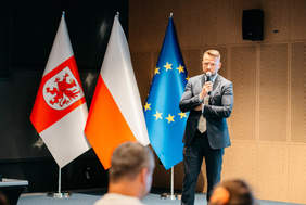 Pokaż zdjęcie: Mężczyzna w ciemnym garniturze i krawacie stoi na scenie, trzymając w prawej ręce mikrofon i przemawiając. Stoi on przed trzema flagami, które są dobrze widoczne: po lewej flaga z czerwonym gryfem na białym tle (prawdopodobnie flaga Województwa Zachodniopomorskiego w Polsce), w środku flaga Polski (biało-czerwona), a po prawej flaga Unii Europejskiej (niebieska z żółtymi gwiazdami). W tle widoczne jest drewniane, panelowe wykończenie ściany. Na pierwszym planie widać tyły głów słuchaczy, siedzących tyłem do obiektywu.