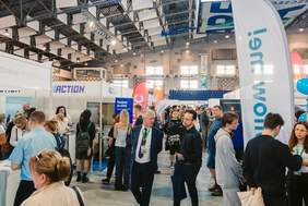 Pokaż zdjęcie: Widok z góry na halę targową podczas wydarzenia w Netto Arenie. Liczne stoiska wystawców oraz odwiedzający rozmawiający przy stanowiskach. W centrum kadru znajduje się baner ‘Demant’ zawieszony pod sufitem. Na pierwszym planie po prawej stronie stoisko ‘Follow me!’ z kolorowymi balonami