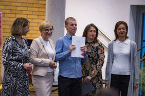 Pokaż zdjęcie: Grupa pięciu osób pozuje do zdjęcia podczas gali konkursowej. W centrum stoi laureat z Dąbrowy Białostockiej, trzymając w dłoniach dyplom. Obok niego znajduje się jego opiekunka oraz trzy kobiety z komisji konkursowej. W tle widoczne są elementy sali i fragmenty prac konkursowych.