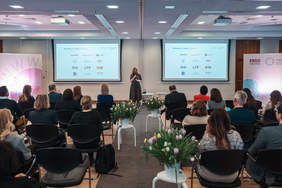 Pokaż zdjęcie: Sala konferencyjna z publicznością siedzącą na krzesłach, na scenie kobieta przemawia do uczestników; dwa ekrany z logotypami partnerów programu, dekoracje z tulipanów na stolikach.