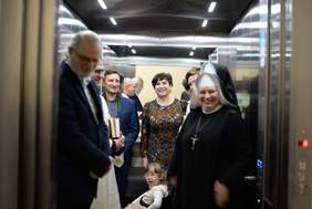 Pokaż zdjęcie: Zdjęcie wnętrza windy, w którym stoi kilka osób.