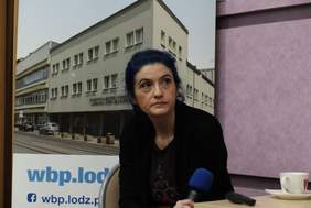 Pokaż zdjęcie: Dr hab. Katarzyna Szmigiero siedzi przy stoliku, trzyma w ręku mikrofon. Po lewej stronie znajduje się zdjęcie biblioteki. 