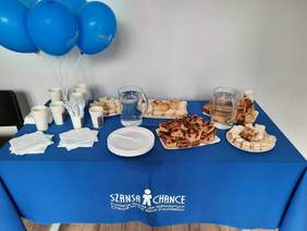 Pokaż zdjęcie: Na stole pokrytym niebieskim obrusem stoi poczęstunek. Są różne rodzaje ciast, woda w dzbankach, talerzyki, sztućce, niebieskie balony. 