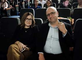 Pokaż zdjęcie: W pierwszym rzędzie siedzą kobieta w czarnych okularach oraz mężczyna w garniturze, uśmiechają się.