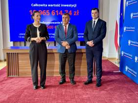 Pokaż zdjęcie: Trzy osoby stoją przed stołem w sali Pomorskiego Urzędu Wojewódzkiego podczas podpisania umowy o dofinansowanie Wspomaganej Społeczności Mieszkaniowej; w tle ekran z kwotą inwestycji 10 965 614,74 zł oraz logotypy Wojewody Pomorskiej i PFRON.