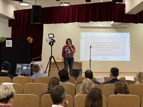 Pokaż zdjęcie: Na scenie stoi kobieta w okularach ubrana w jeansy i kolorowy sweter. W ręku trzyma mikrofon. Po lewej stronie stoi aparat na statywie transmitujący wykład online.