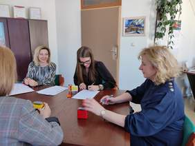 Pokaż zdjęcie: W gabinecie przy ciemnym okrągłym stole siedzą cztery kobiety elegancko ubrane. Na stole leżą dokumenty oraz stoi pieczątka. Kobieta druga od prawej, w okularach z ciemnymi długimi włosami podpisuje umowę.