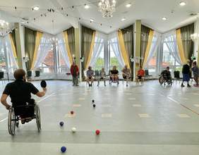 Pokaż zdjęcie: W dużej sali znajdują się trzy osoby poruszające się na wózku, z czego jedna z nich trzyma w ręce paletkę, mając swój wzrok skierowany na grupę dziewięciu osób znajdujących się pod oknem. Na podłodze rozłożone są różnokolorowe małe piłki.