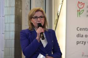 Pokaż zdjęcie: Kobieta blondynka w okularach i niebieskim garniturze, stoi z mikrofonem i przemawia. W lewej ręce trzyma dokumenty. W tle widać fragment baneru z logo PFRON