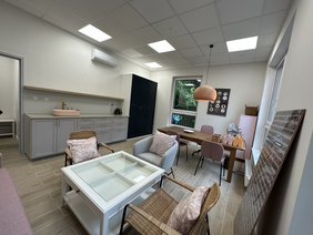 Pokaż zdjęcie: W Sali kuchnia z umywalką i czarną szafa. Obok okno stół drewniany na cztery osoby, na pierwszym planie stół z 4 przegródkami pod szkłem i dwoma fotelami.