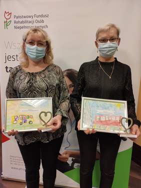 Pokaż zdjęcie: dwie kobiety w okularach stoją na tle banneru PFRON w rękach trzymają kolorowe obrazki narysowane przez dzieci i statuetki w kształcie serca