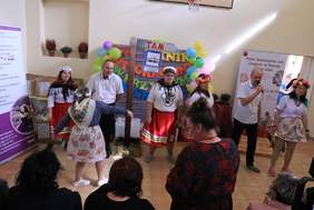 Pokaż zdjęcie: Zdjęcie przedstawia występ artystyczny uczestników WTZ. Impreza integracyjna odbywa się w sali gimnastycznej, grupa osób śpiewa i tańczy . Panowie ubrani w białą koszule i czarne spodnie, Panie w kolorowe spódnice i białe bluzki na głowie maj